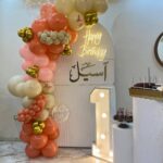 تنسيق خلفية محفورة بيضاء مع إضاءة نيون Happy Birthday واسم أسيل ورقم 1 مضيء وبالونات مشمشي وأوف وايت وذهبي مع فراشات بجدة - متجر زينة بارتي