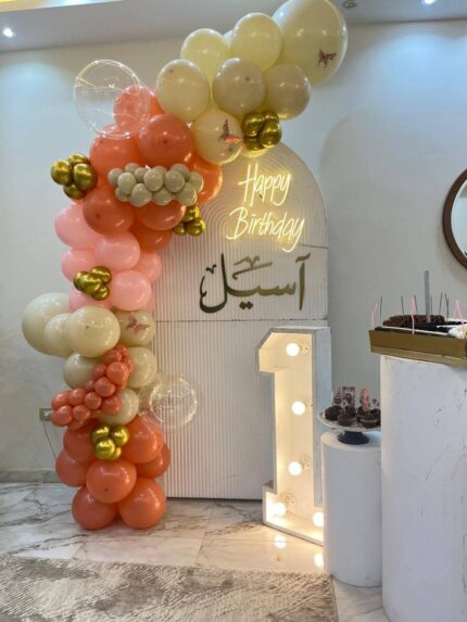 تنسيق خلفية محفورة بيضاء مع إضاءة نيون Happy Birthday واسم أسيل ورقم 1 مضيء وبالونات مشمشي وأوف وايت وذهبي مع فراشات بجدة - متجر زينة بارتي